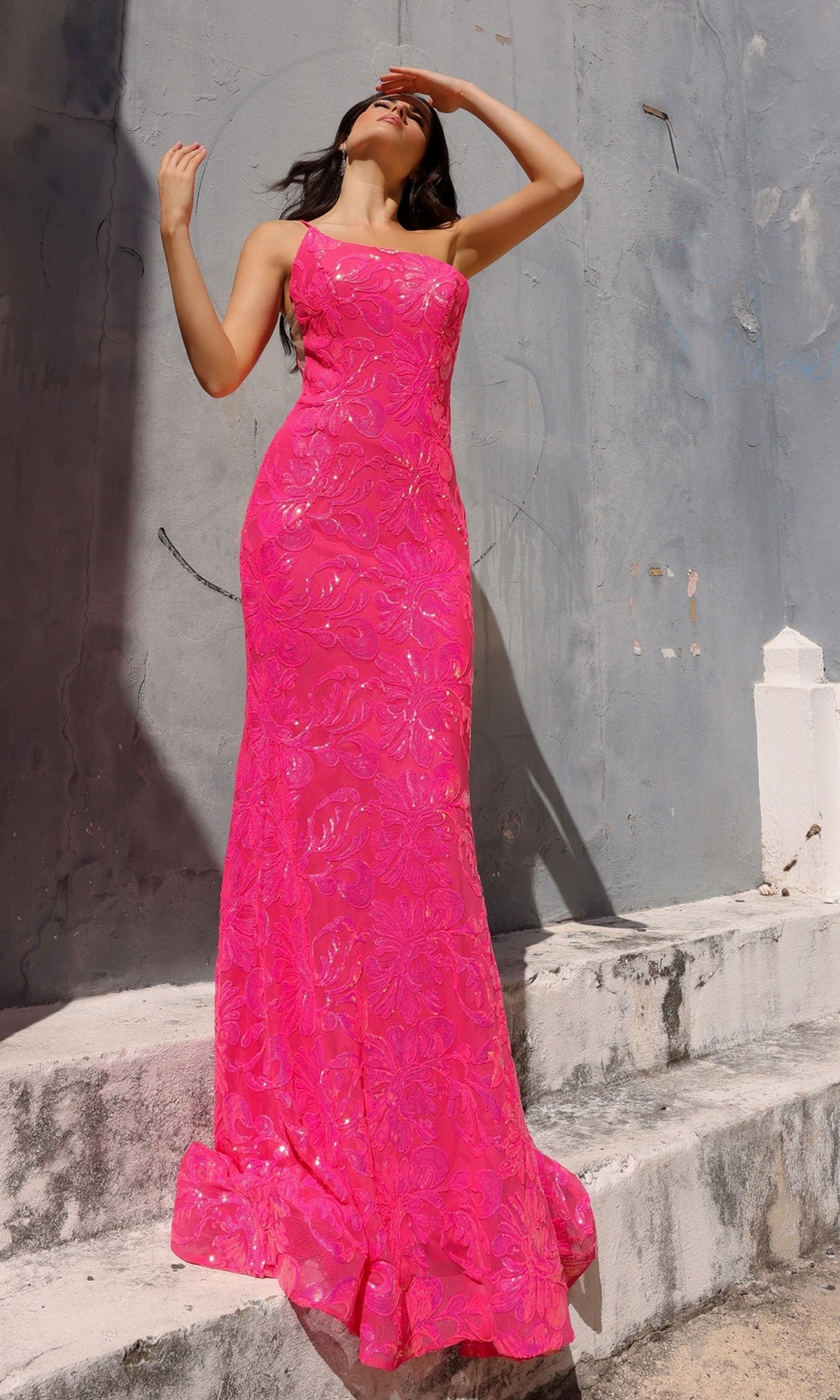 A woman in the One-Shoulder Long Sequin-Print Prom Dress R1308 stands on stone steps by a weathered gray wall, raising one hand to shield her face from the sun.